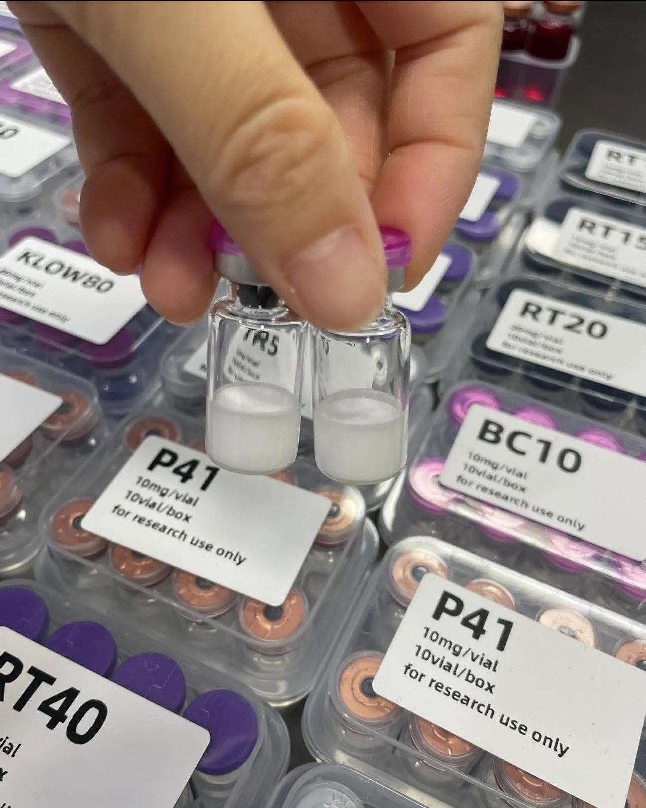 Two research peptide vials held for inspection above inventory trays.